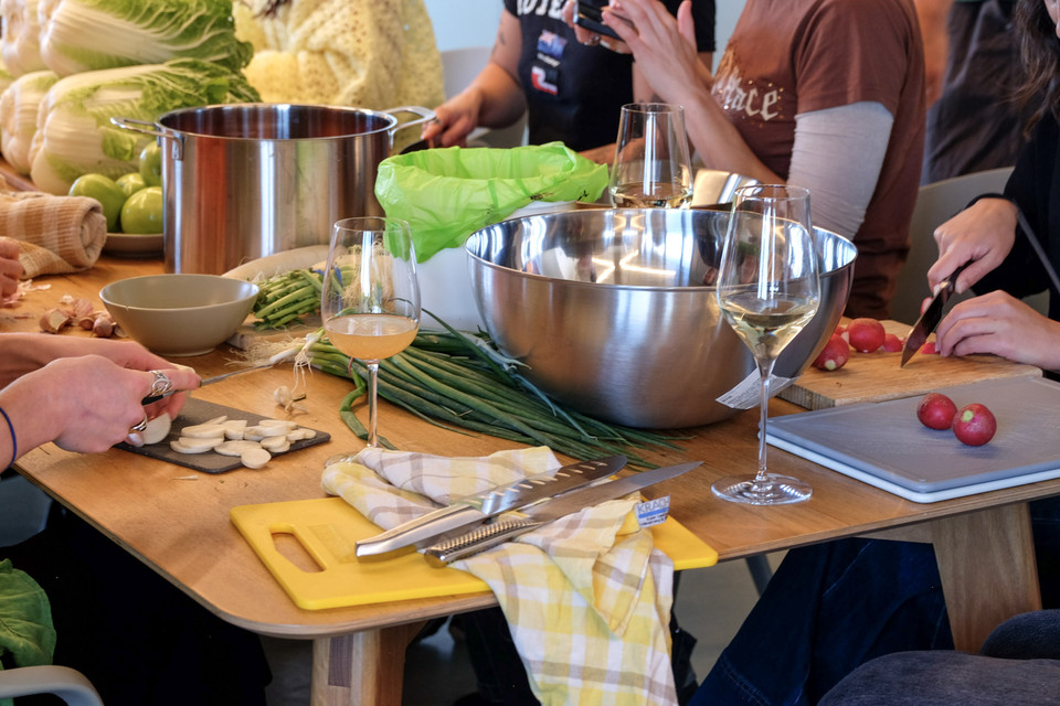 Hands slice garlic and radishes on boards around a big steel bowl and a bundle of spring onions on a wooden table.