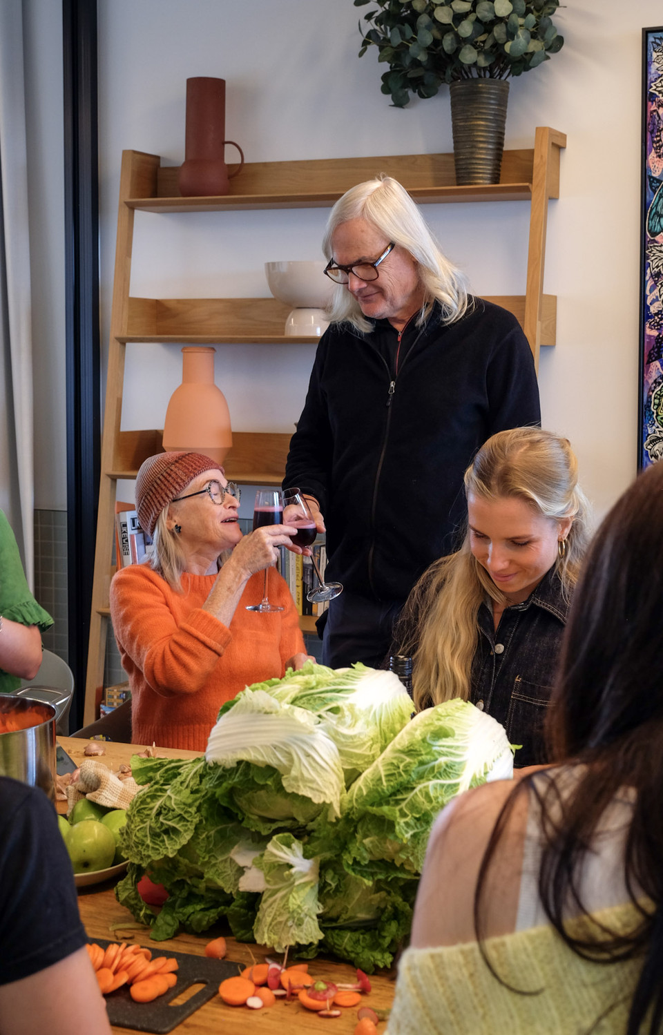 An older woman in an orange jumper raises a glass of red wine beside a pile of wombok as friends gather round.