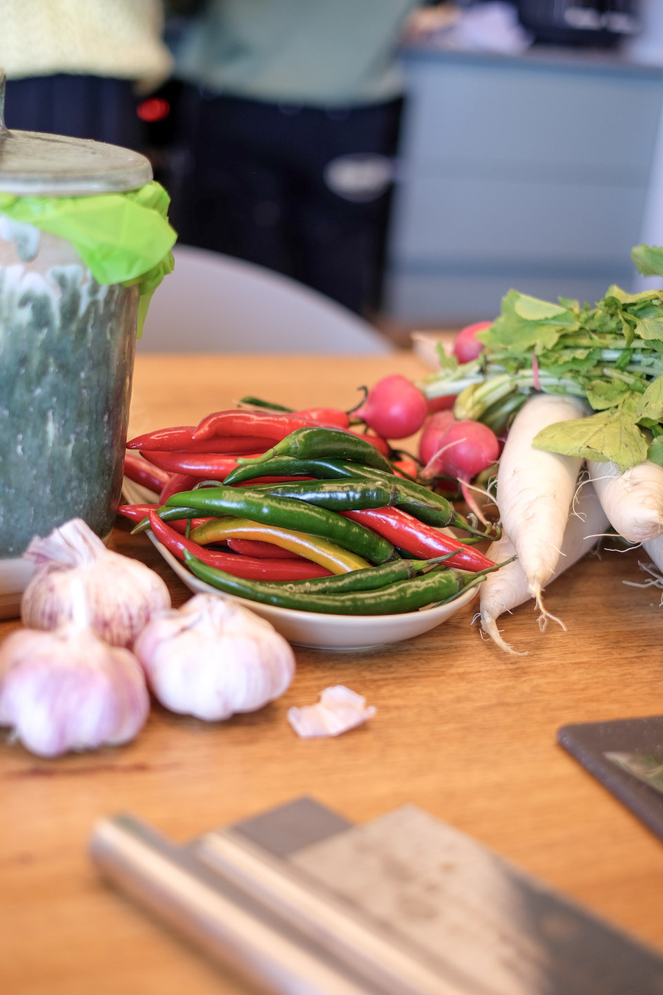 A plate of red and green chillies sits beside garlic bulbs, radishes and daikon on a wooden table.