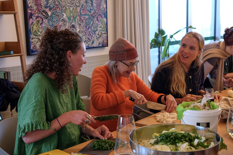 Three people laugh together while chopping greens at a long table in a sunlit room.