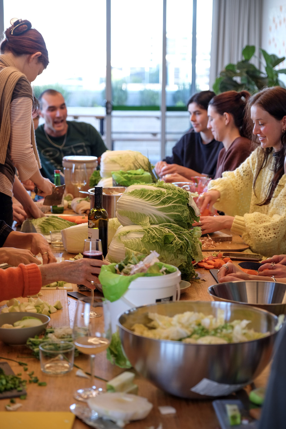 People chop vegetables and laugh around a long table stacked with whole cabbages.