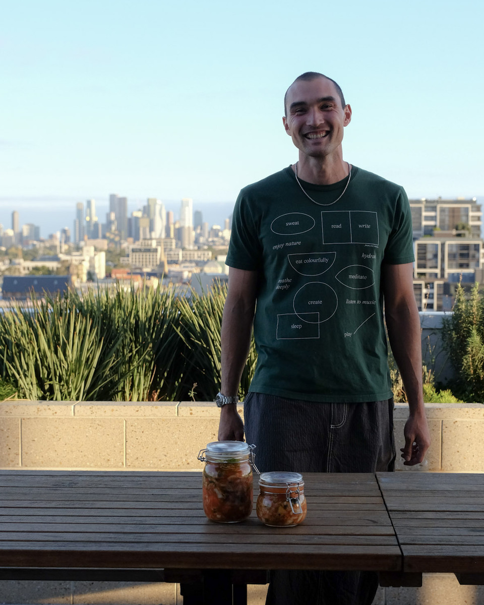 A smiling man in a green t-shirt stands behind a wooden table with two jars of kimchi, the city skyline behind him.