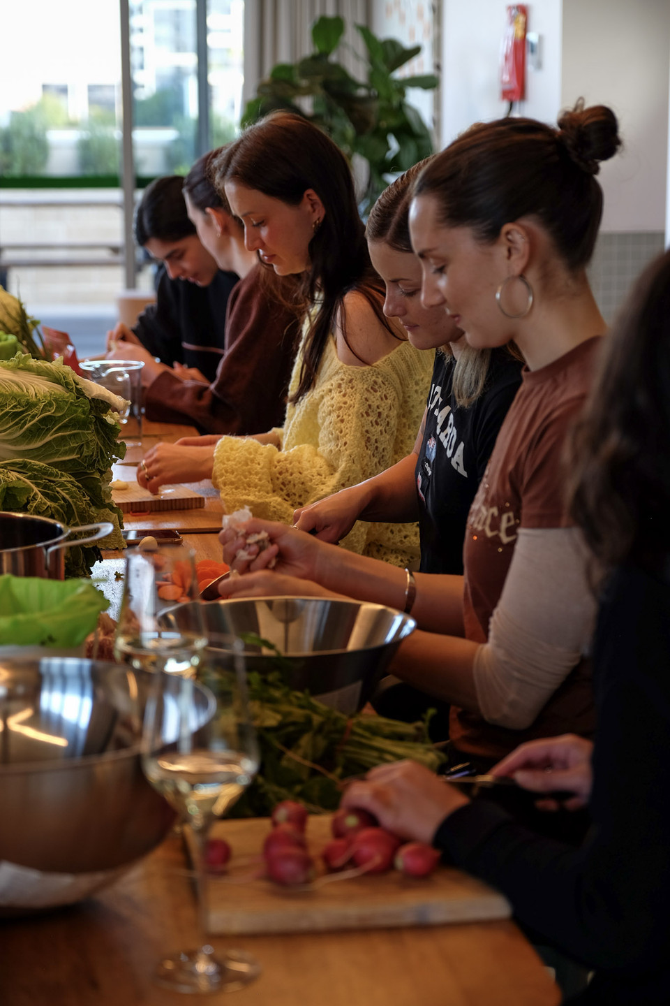 A row of people look down as they chop radishes and greens along a long wooden table in a bright room.