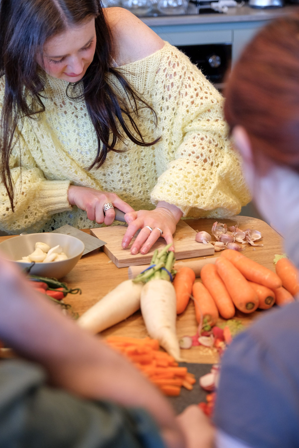 A woman in a yellow knit jumper slices garlic on a board, with daikon, carrots and a bowl of cloves on the table.