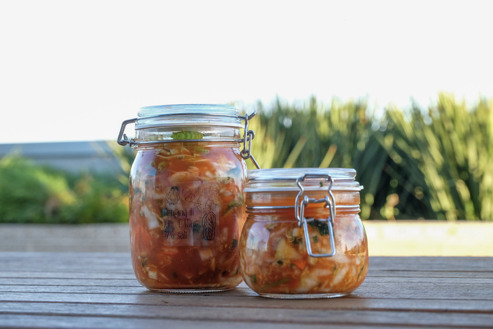 Two clip-top jars of kimchi sit on a weathered outdoor table, with green shrubs and open sky softly behind.