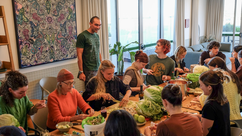 A bright room of people chopping vegetables along one long table, with a tall man standing to talk them through it.