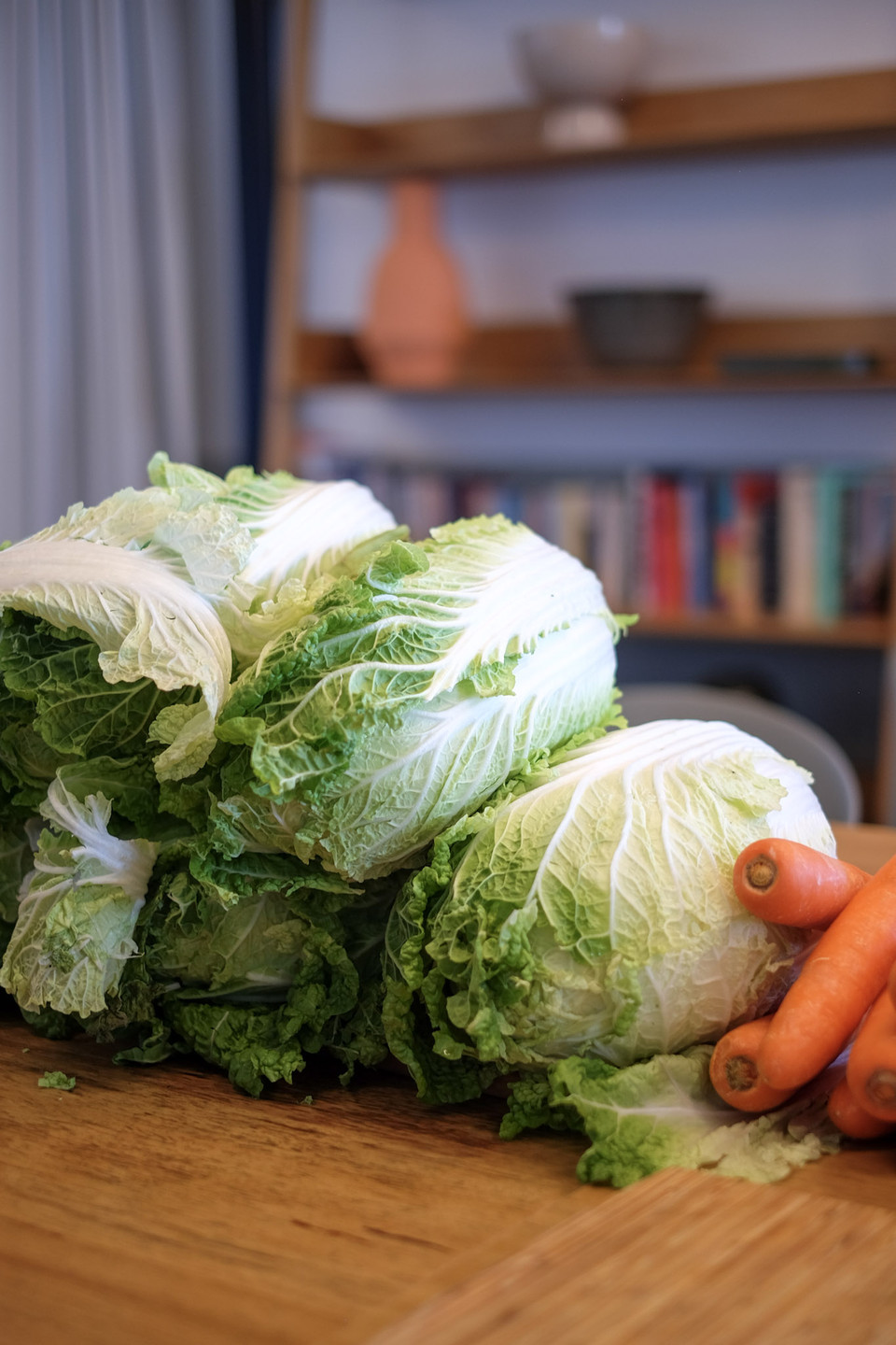 Whole wombok cabbages sit on a wooden table beside a handful of carrots, with a shelf of books softly behind.